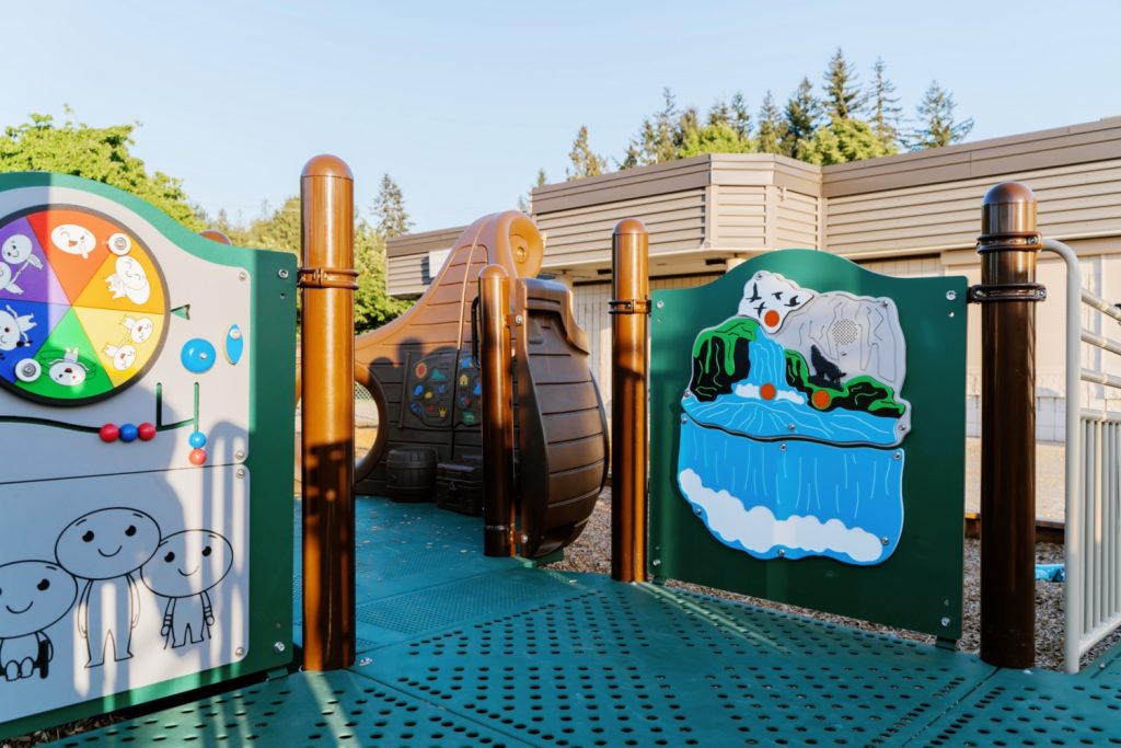 École Centennial Park Elementary-03 Playground at École Centennial Park Elementary with a colorful activity panel displaying a wheel and smiling stick figures. A nearby rocker resembles a pirate ship, surrounded by trees.