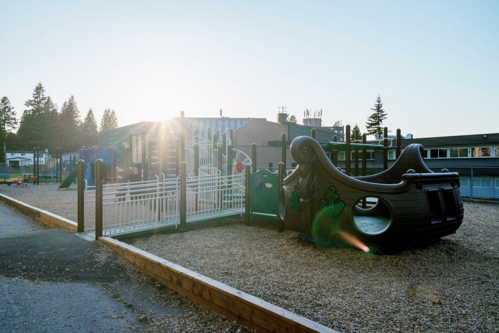 École Centennial Park Elementary-09 Playground at École Centennial Park Elementary with a pirate ship rocker and slides, bathed in morning sunlight. Trees and a building are visible in the background. Calm and inviting atmosphere.
