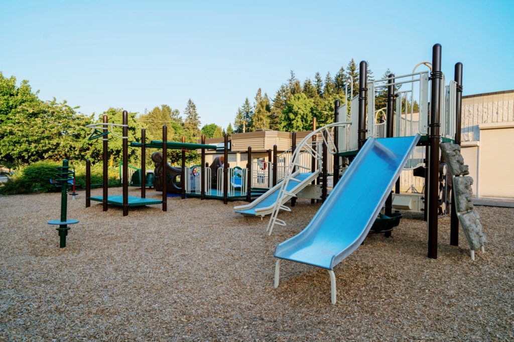 École Centennial Park Elementary-10 Playground set with slides and climbing structures on wood chips, surrounded by trees under a clear blue sky, conveying a peaceful, inviting atmosphere.