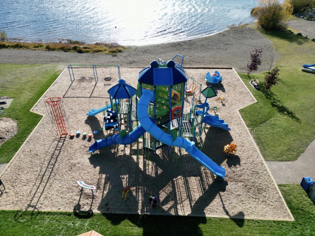 Radley Beach Playground-01 Aerial view of Radley Beach Playground by a lake. Features include blue slides, climbing structures, and swings on a wood chip area. Sunlight sparkles on the water nearby.