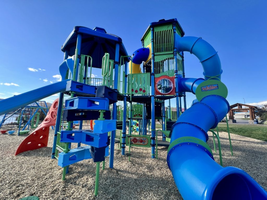 Radley Beach Playground-05 Radley Beach Playground with vibrant blue and green structures under a clear sky. Includes slides, climbing frames, and tunnels, conveying fun and adventure.