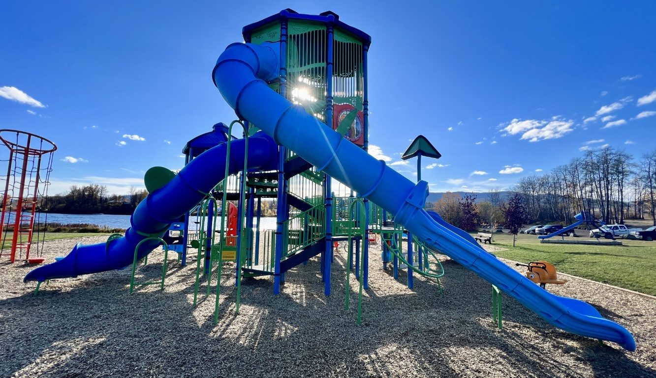 A tall, colorful playground structure at Radley Beach Playground with bright blue tube slides sits under a clear blue sky. The area is lively and sunlit, with trees in the background.