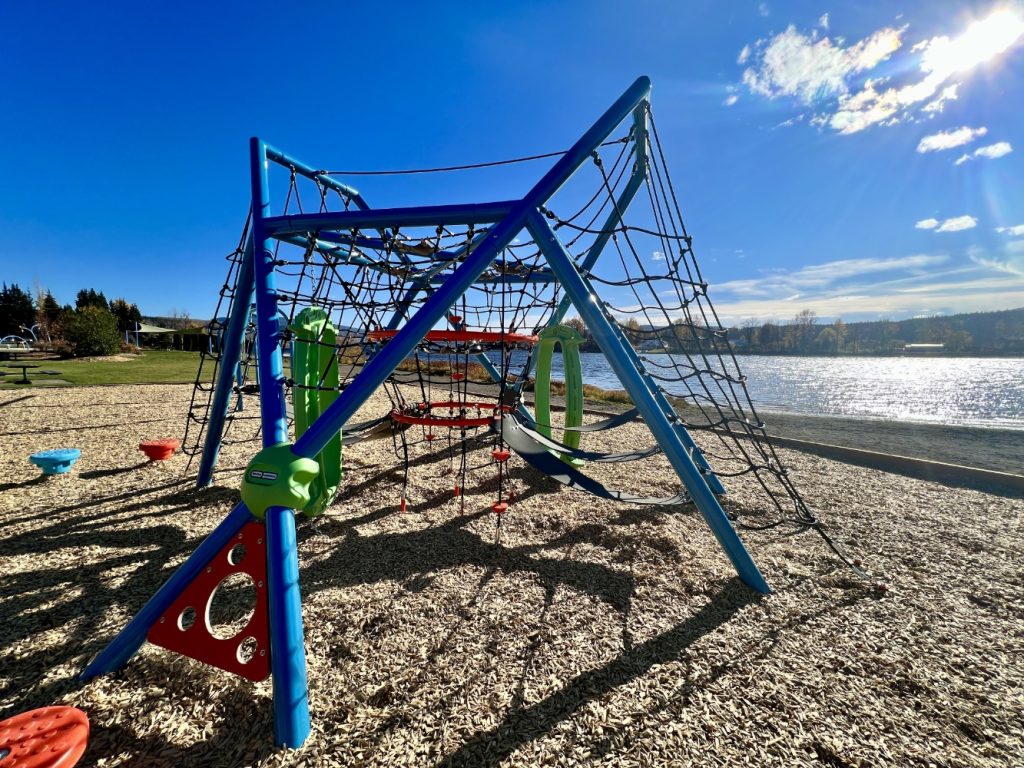 Radley Beach Playground-09 Radley Beach Playground with a net climbing structure by a sunny lakeside, with wood chip ground and vibrant blue sky, creating a joyful atmosphere.
