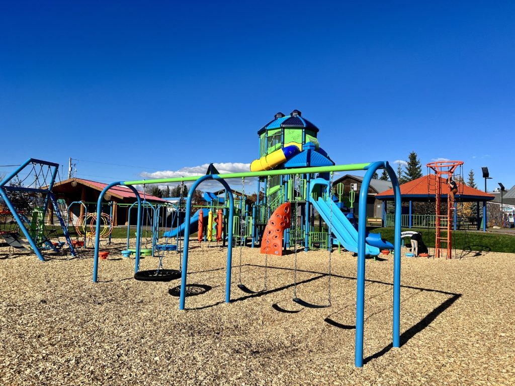 Radley Beach Playground-10 Playground under a clear blue sky, featuring colorful slides, swings, climbing frames, and a wood chip ground, creating a joyful and vibrant atmosphere.