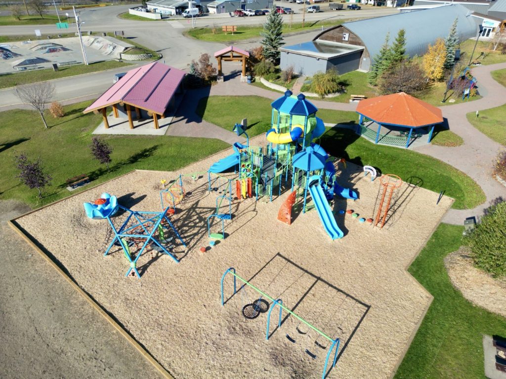 Radley Beach Playground-11 A bird's-eye view of Radley Beach Playground with slides, climbing structures, and swings on wood chips. Nearby, a green lawn hosts picnic shelters. Bright and inviting.