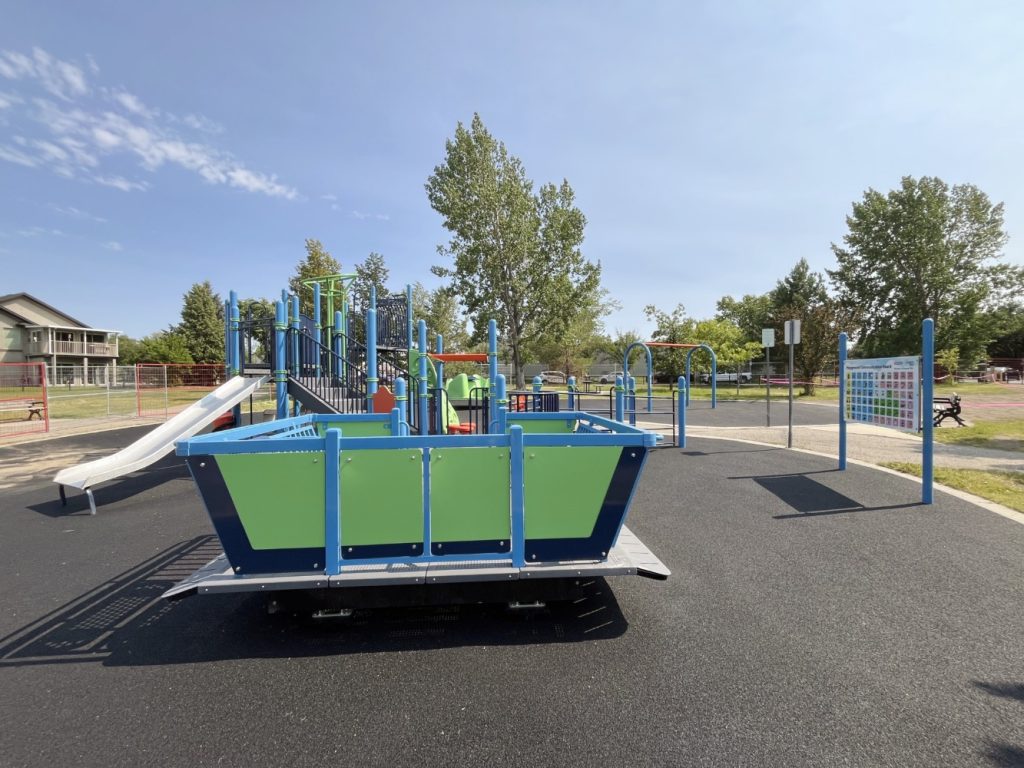 Chinook Lake Park-02 Inclusive playground with a wheelchair-accessible rocker, colorful slides, and climbing structures. Surrounded by trees under a clear blue sky.