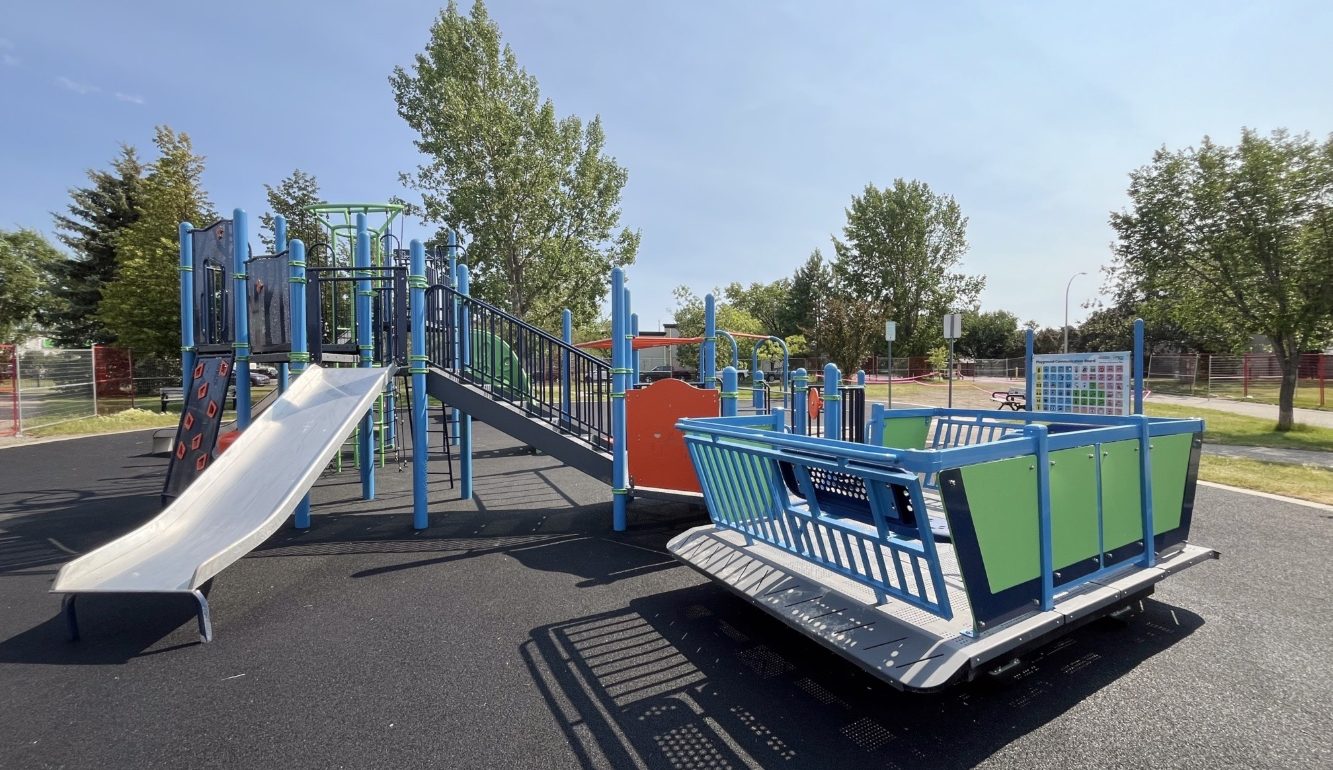 Playground at Chinook Lake Park with blue and green equipment under a clear sky. Features slides, climbing structures, and trees in the background, conveying a cheerful atmosphere.