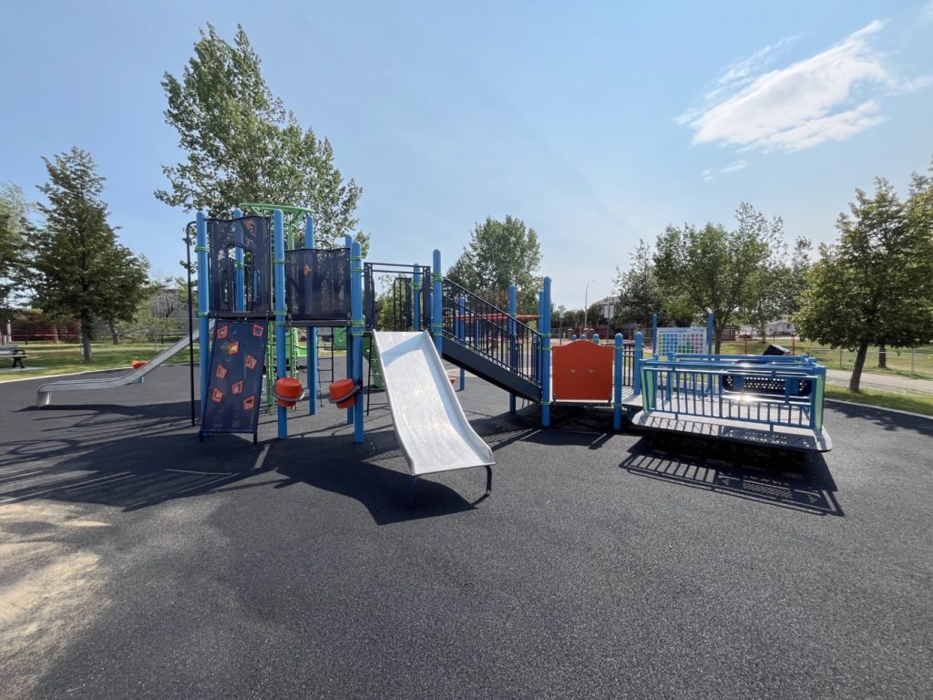 Chinook Lake Park-04 Playground under clear sky, featuring stainless steel slides, blue and orange climbing structures, and swings over soft black flooring, surrounded by trees.