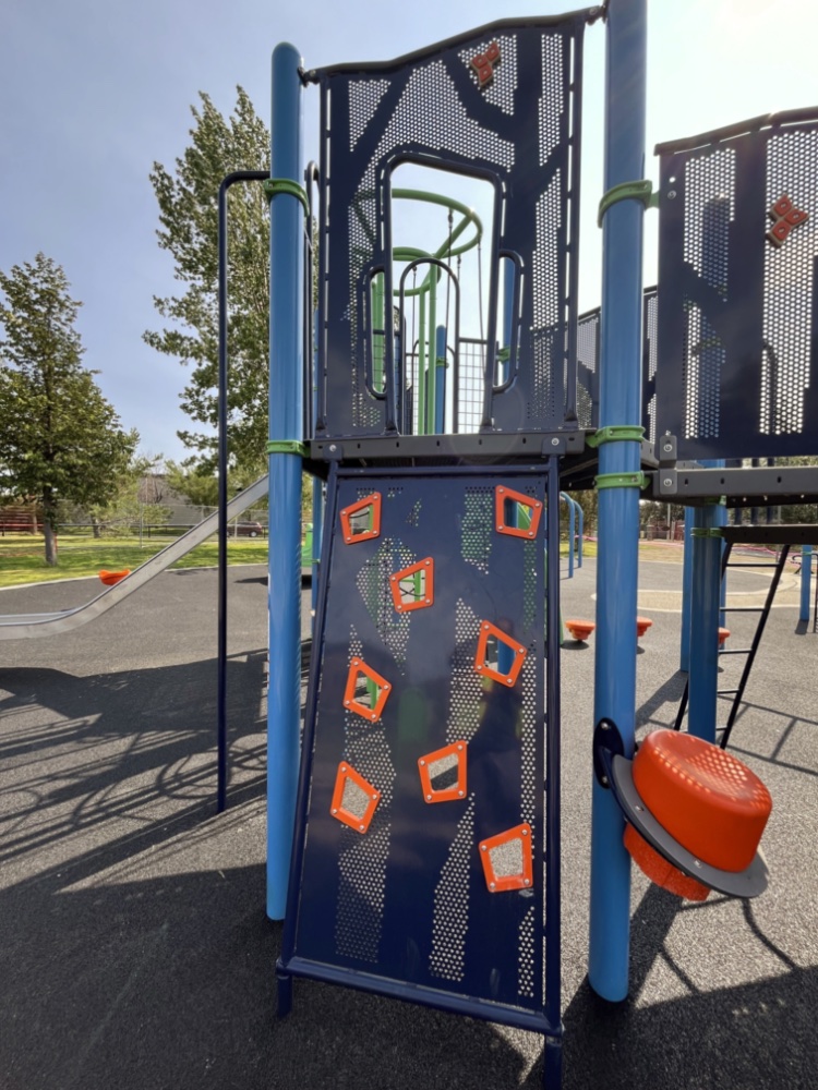 Chinook Lake Park-05 Playground at Chinook Lake Park with a climbing wall featuring orange handholds, blue posts, and a sliding pole. Trees and a clear sky suggest a bright, sunny day.