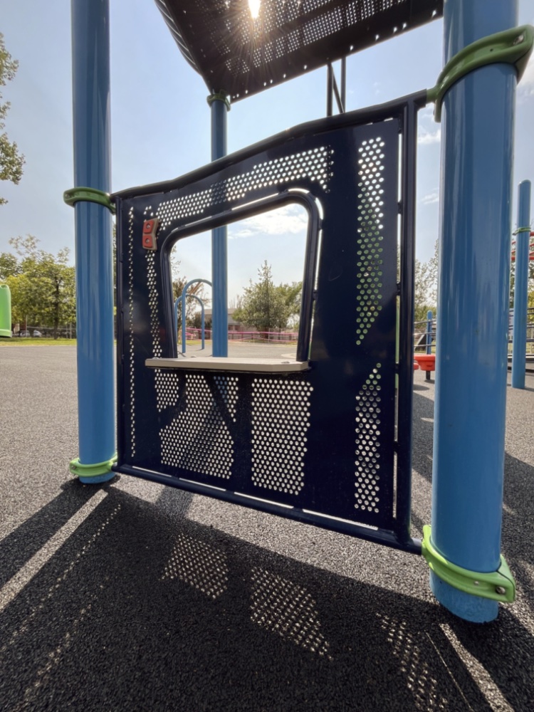 Chinook Lake Park-06 Blue playground activity panel with perforated metal in sunlight, casting shadows on the ground. Green elements provide contrast, evoking a playful mood.