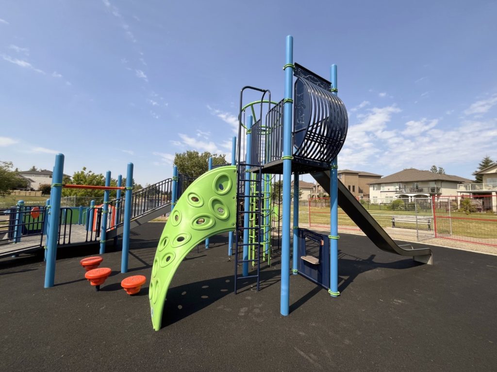 Chinook Lake Park-09 Colorful playground at Chinook Lake Park featuring a tall blue and green climbing structure with a slide, located on a sunny day surrounded by houses and trees.
