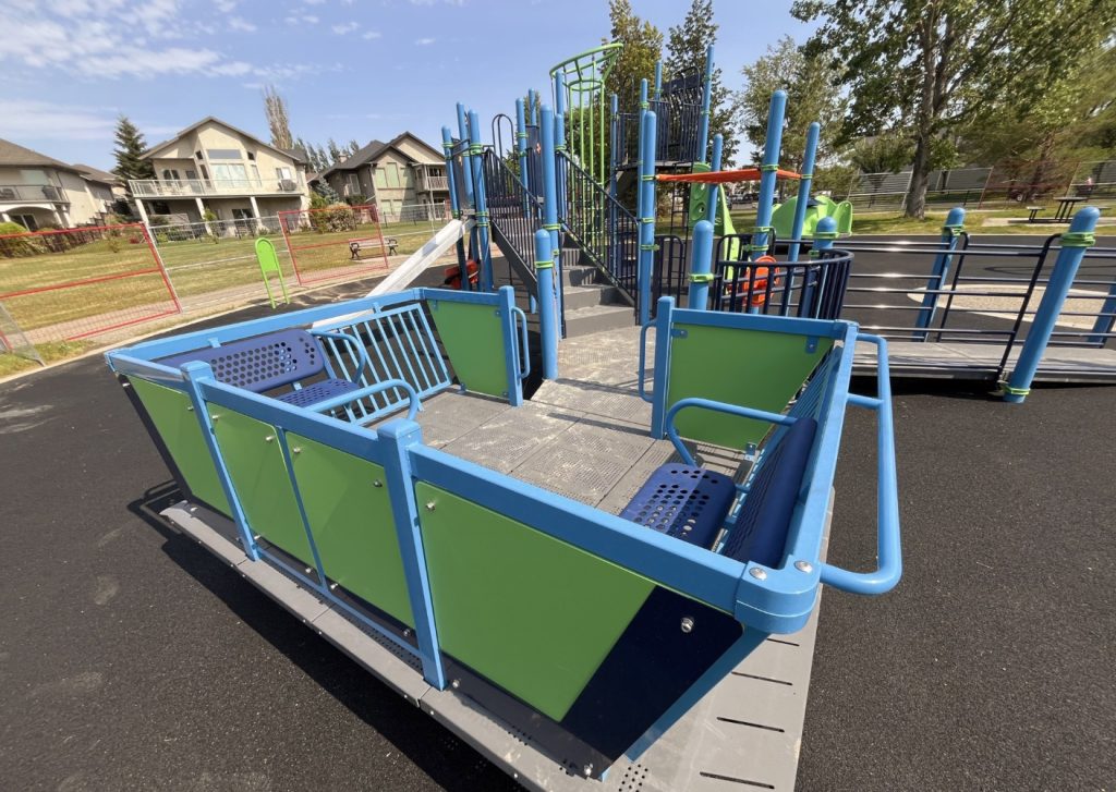 Chinook Lake Park-12 Inclusive playground with green and blue wheelchair-accessible rocker, including ramps and platforms. Surrounded by grassy area and houses. Clear, sunny day.