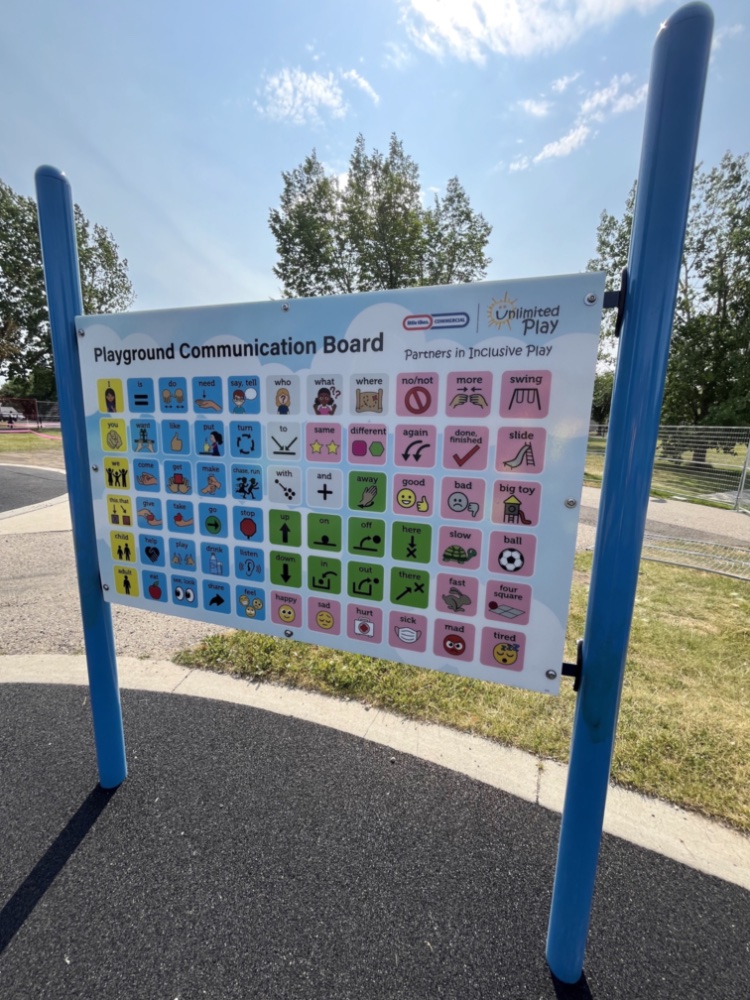 Chinook Lake Park-13 Playground communication board at Chinook Lake Park with colorful symbols and text for inclusive play, mounted on blue poles. Sunny day with trees in the background.