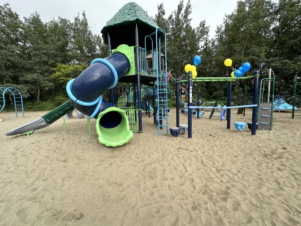 École Sacré Coeur-01 Bright playground at École Sacré Coeur with green and blue slides, climbing structures, and yellow balloons. Set on sand with surrounding trees, conveying a playful atmosphere.