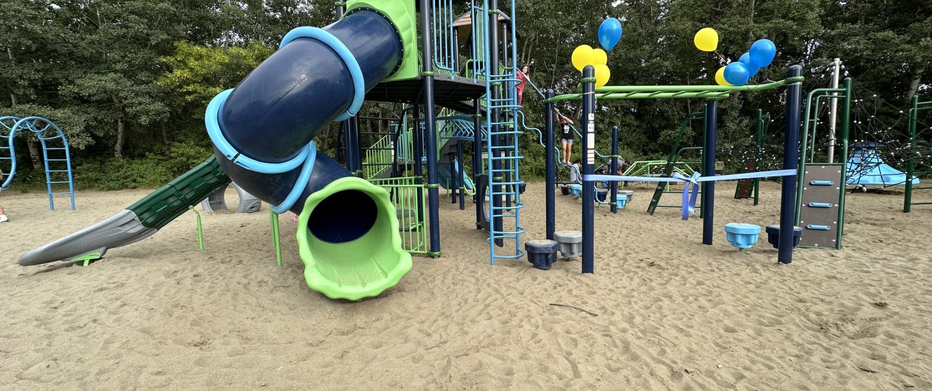 Bright playground at École Sacré Coeur with green and blue slides, climbing structures, and yellow balloons. Set on sand with surrounding trees, conveying a playful atmosphere.