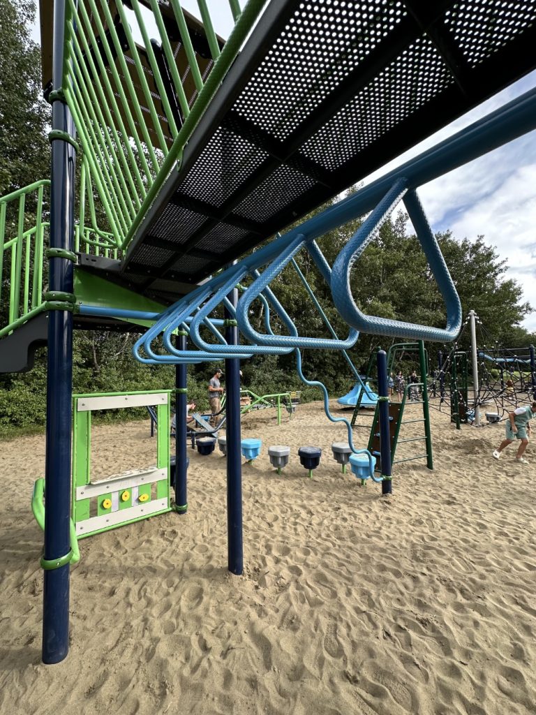 École Sacré Coeur-10 Playground with blue monkey bars and a bright green structure in the foreground. Children are playing in the background, creating a lively atmosphere. Trees border the area, sky is partly cloudy.