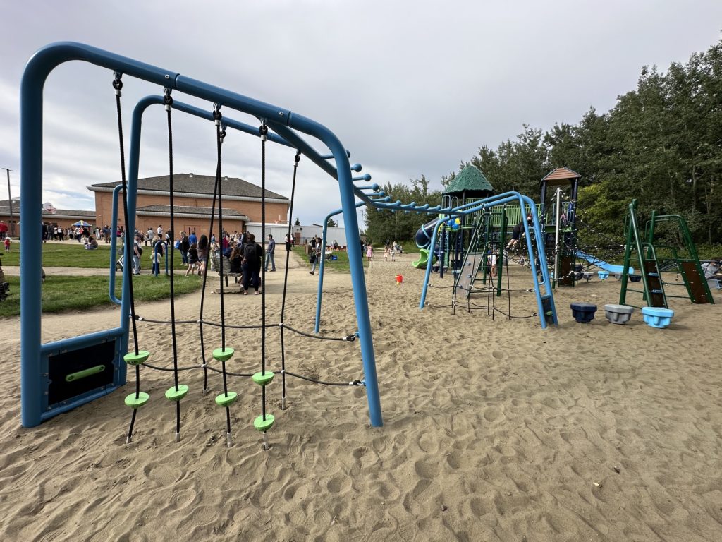 École Sacré Coeur-12 Playground with blue climbing structures and ropes on sand, surrounded by trees and a school building. Families and children play on a cloudy day.