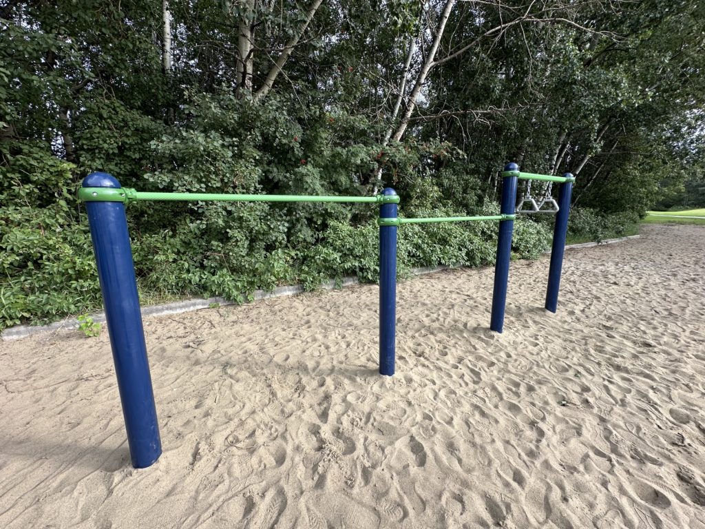 École Sacré Coeur-15 Blue and green pull-up bars are set on a sandy playground at École Sacré Coeur with lush green trees in the background, creating a serene outdoor workout space.