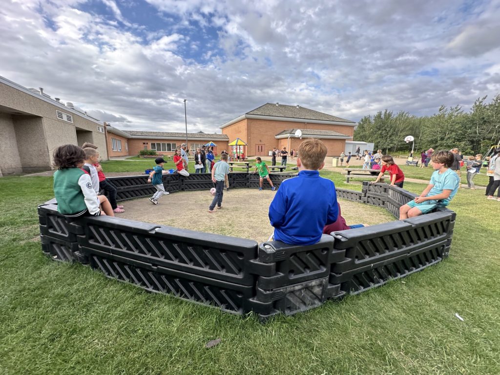 École Sacré Coeur-16 Children play with a soccer ball in a gaga ball pit on grass, surrounded by school buildings and trees under a cloudy sky. The scene is lively and joyful.