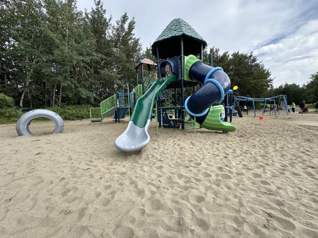 École Sacré Coeur-02 Playground scene with a large, colorful play structure featuring a twisting slide and climbing areas. Surrounded by sand and trees, creating a playful, inviting atmosphere.