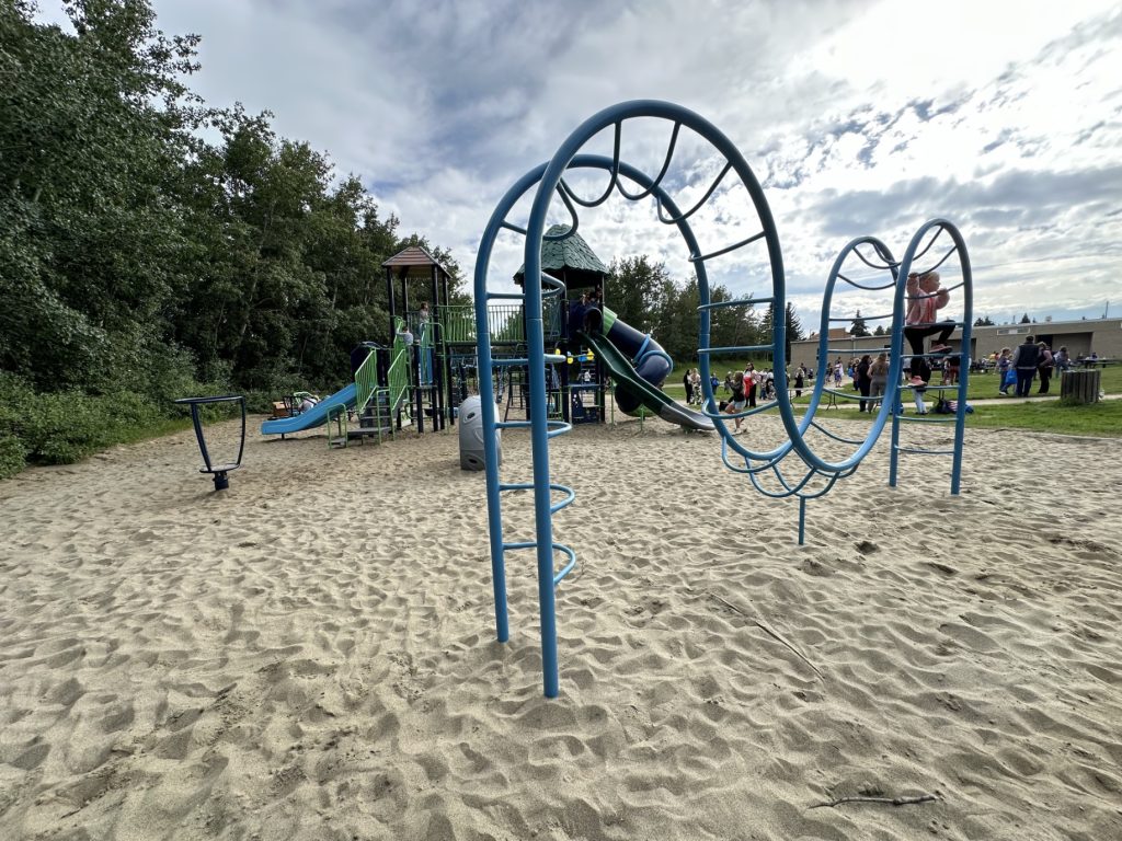 École Sacré Coeur-03 Playground at École Sacré Coeur with blue climbing frame, slides, and children playing on sandy ground. Trees and a gathering of people in the distance under a cloudy sky.