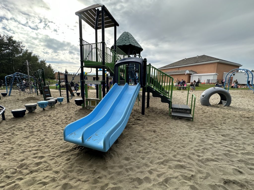 École Sacré Coeur-06 Playground scene with a blue double slide and green climbing structure on sandy ground. Children and parents are gathered in the background under cloudy skies.