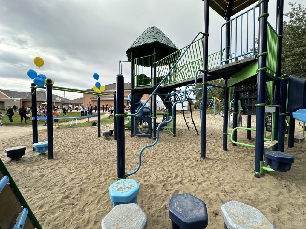 École Sacré Coeur-08 Playground with green, blue, and grey equipment on sand. Balloons float nearby, and people gather in the background on grass, creating a cheerful atmosphere.