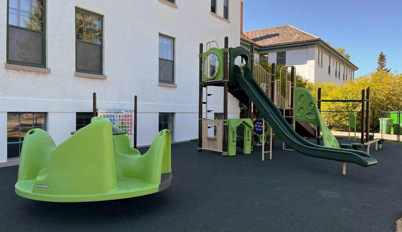 A playground at New Heights School with green equipment, including a slide and merry-go-round, sits on a dark rubber surface beside a white building under a clear blue sky.