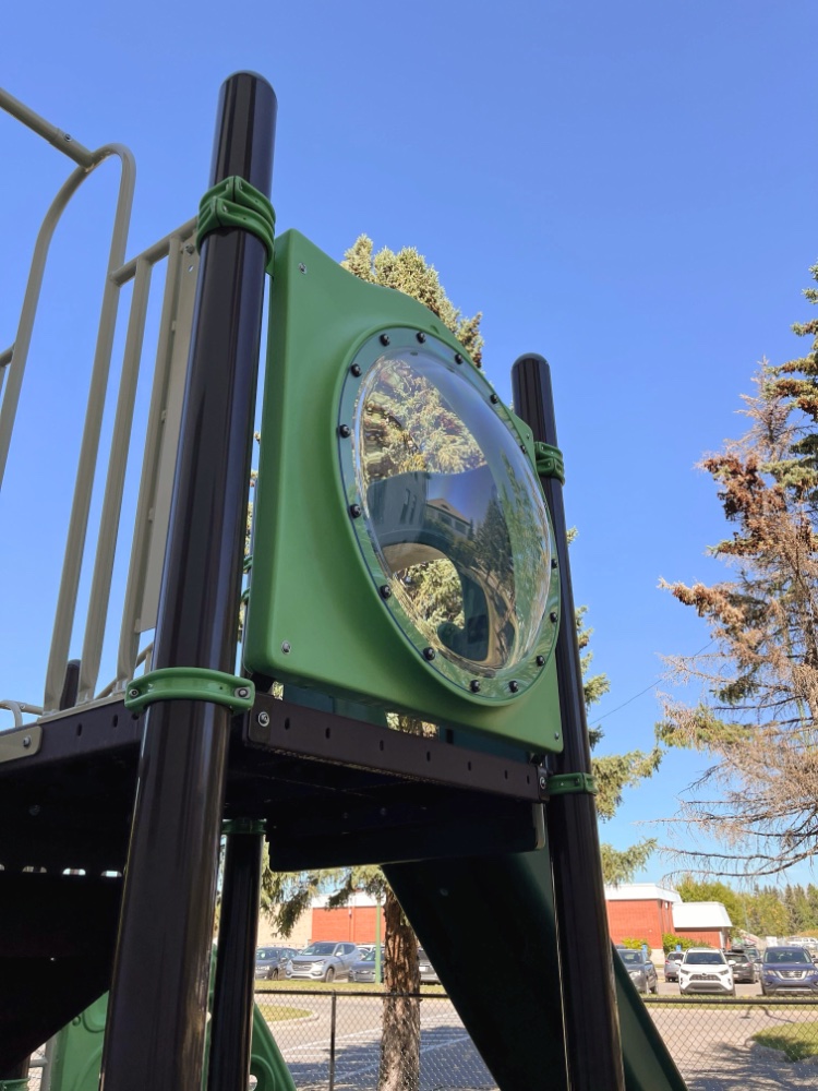 New Heights School-02 Playground climbing structure with brown and green components, featuring a circular bubble window reflecting trees. Clear blue sky in the background.
