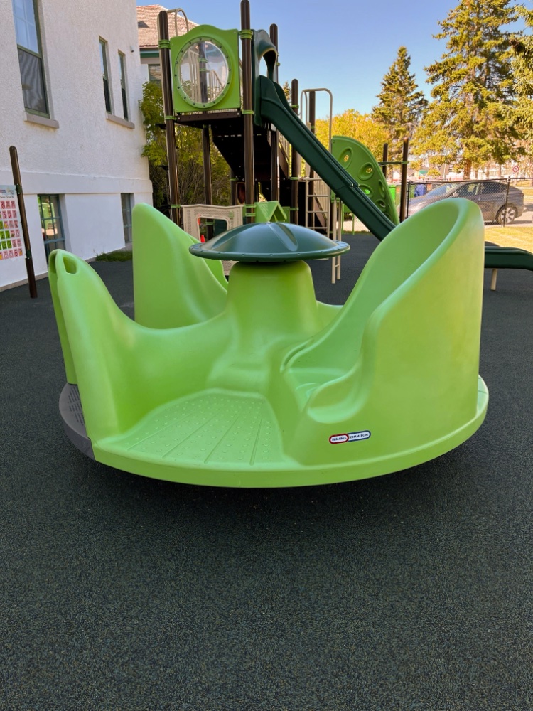 New Heights School-03 A light green playground spinner at New Heights School stands on a soft black surface. In the background, there's a slide and a climbing structure next to a building, with trees nearby.