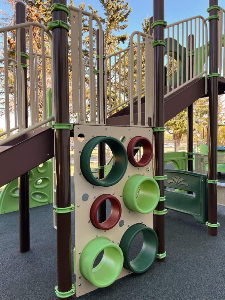 New Heights School-06 Playground structure with a climbing panel featuring circular cutouts in burgundy and green. Stairs and slides visible, set in a park with trees. Playful mood.