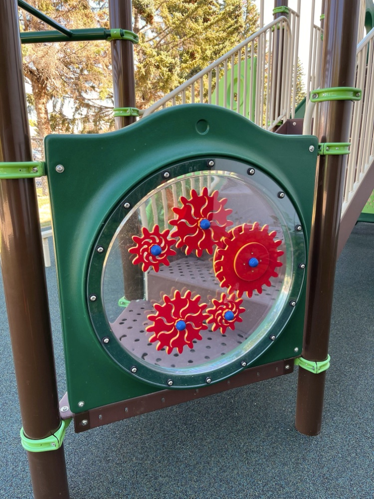 New Heights School-07 Playground panel at New Heights School with four interlocking red gears inside a clear, round window. Set against a green frame. Background shows trees and equipment.