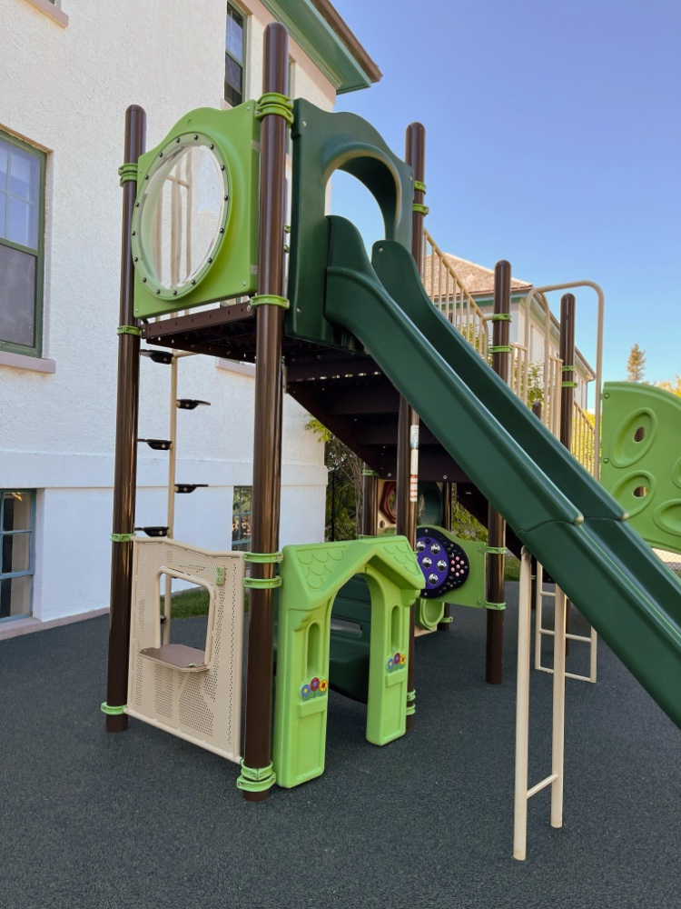New Heights School-09 A bright playground with green slides, climbing structures, and playhouse, set on a soft, dark ground. The scene feels inviting and playful.