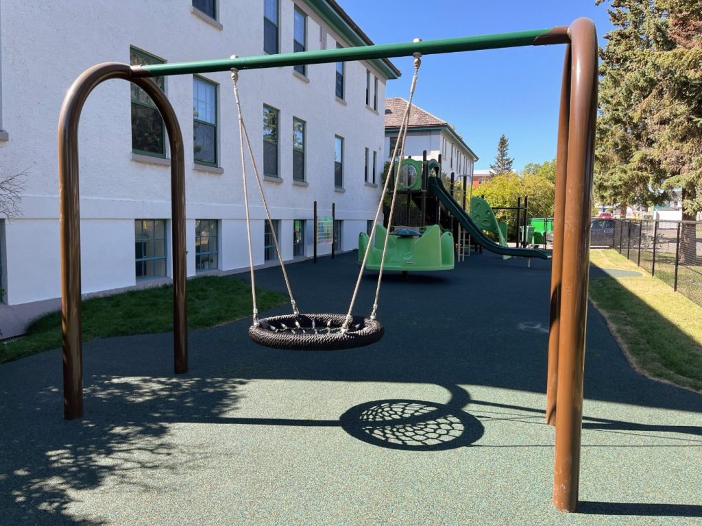 New Heights School-11 Playground at New Heights School with circular swing in the foreground, casting a shadow. Slide and climbing structure in the background, next to a two-story building. Sunny day.