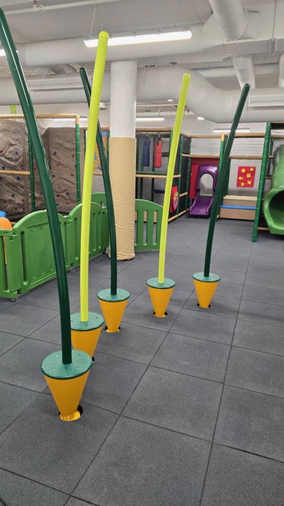 Village Square Leisure Centre-10 Indoor playground with grey padded floors. Tall, flexible poles rise from cone bases, surrounded by soft barriers, climbing walls, and slides. Bright, playful vibe.