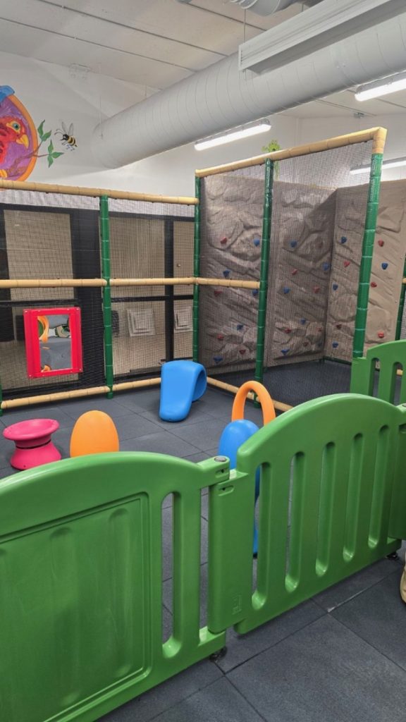 Village Square Leisure Centre-11 Indoor playground at Village Square Leisure Centre with a green fence, colorful play equipment, and rock climbing walls. Bright, playful atmosphere with cartoon wall art.