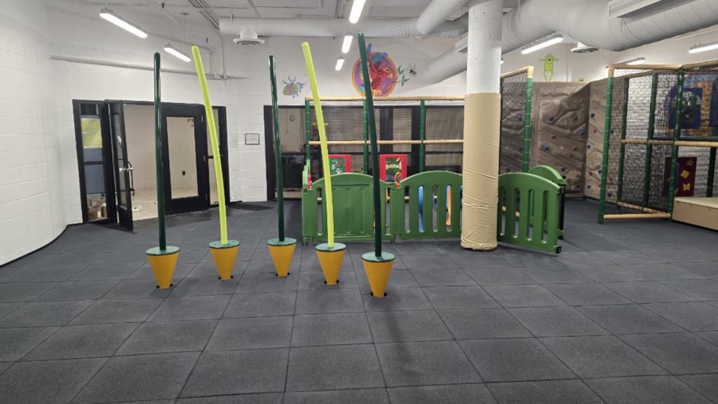 Village Square Leisure Centre-03 Indoor playground at Village Square Leisure Centre with padded flooring, featuring tall green and yellow poles resembling trees. A climbing wall is visible in the background. The mood is playful and safe.