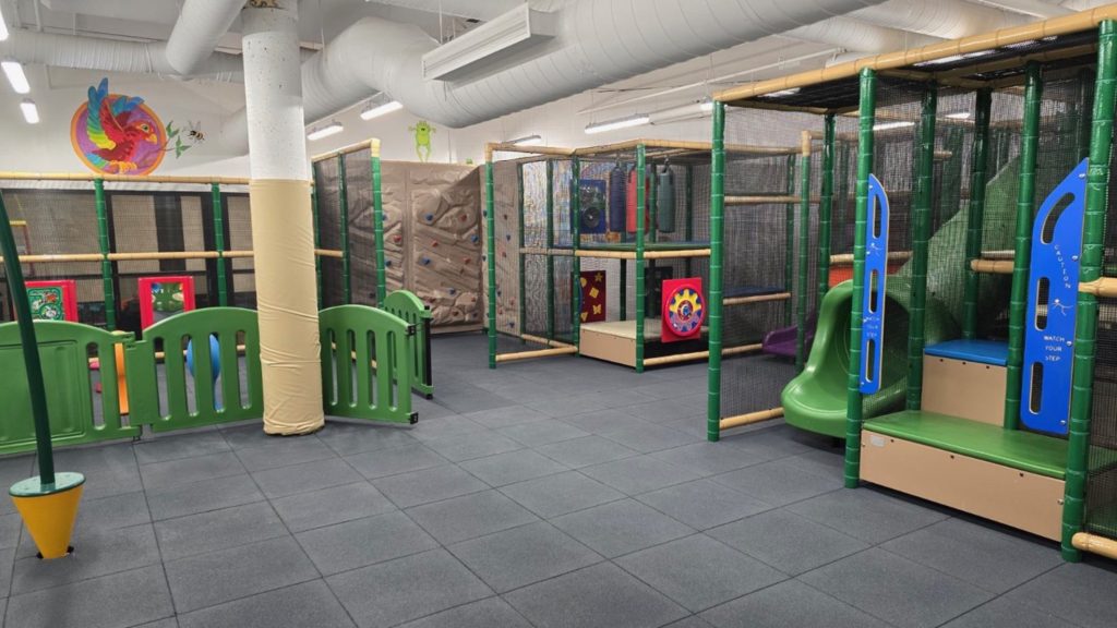 Village Square Leisure Centre-04 Indoor play area with climbing structures, slides, and colorful panels. Rubber flooring for safety. Bright, cheerful atmosphere with playful designs.