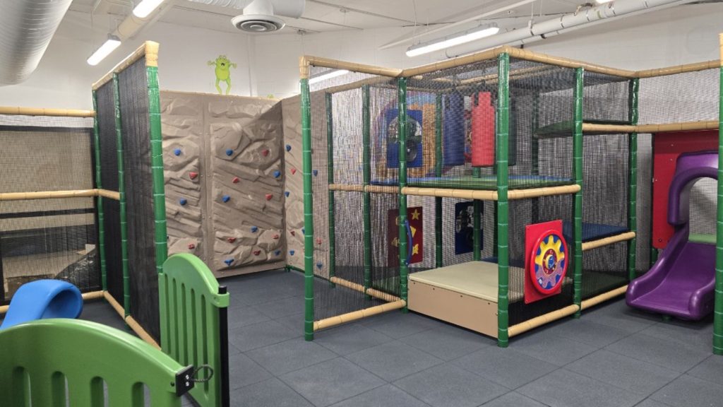 Village Square Leisure Centre-07 Indoor playground at Village Square Leisure Centre with climbing wall, purple slide, and soft play structures. Enclosed by netting and framed with green and beige poles. Bright, playful atmosphere.
