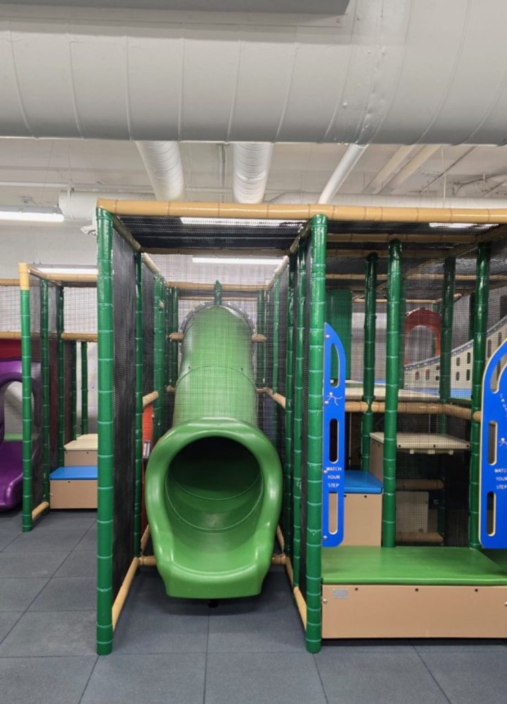 Village Square Leisure Centre-08 Indoor playground featuring a green tunnel slide surrounded by netted climbing structures and platforms. Bright, fun atmosphere with beige and blue accents.