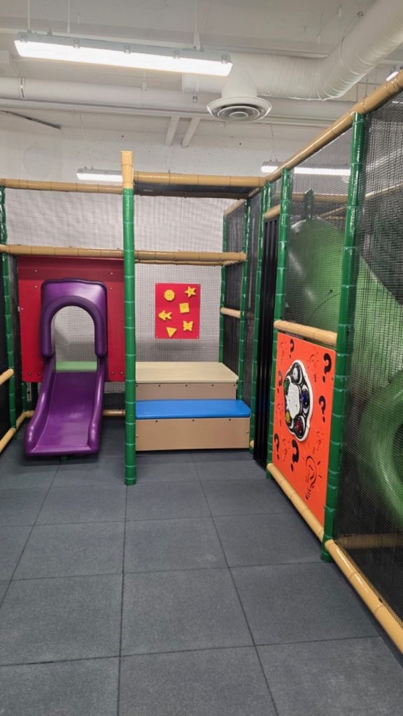 Village Square Leisure Centre-09 Indoor playground at Village Square Leisure Centre with padded flooring, featuring a purple slide, climbing steps, and colorful wall panels with shapes and patterns. Bright, playful atmosphere.