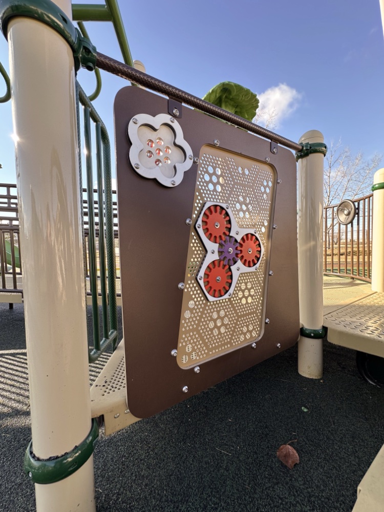 Willow Park School-05 Playground equipment at Willow Park School with a panel of rotating red and purple gears on brown and beige frame. Clear sky and trees in background. Energetic, playful scene.