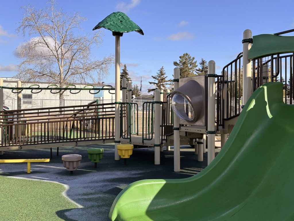 Willow Park School-06 Playground with a green slide, climbing structures, and a bridge under a blue sky. Trees and a building are in the background, creating a playful and inviting atmosphere.
