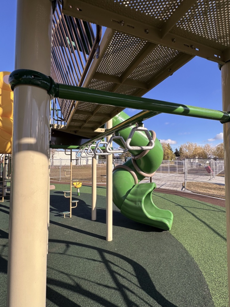 Willow Park School-12 Playground with green spiral slide, beige climbing structures, and monkey bars. Sunny day, blue sky, and a few clouds create a cheerful atmosphere.