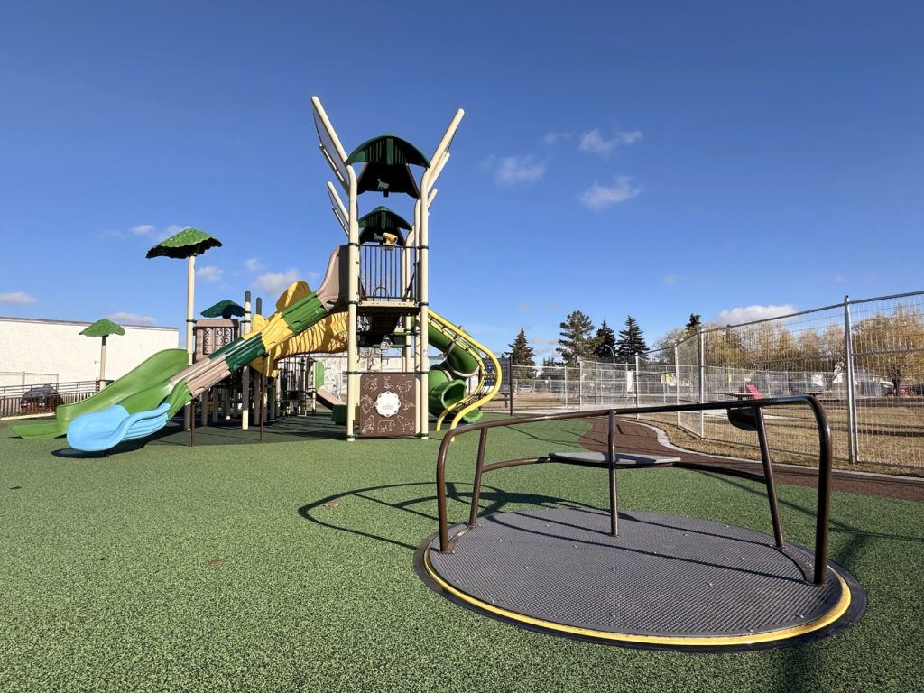 Willow Park School-13 Playground at Willow Park School with a treehouse-themed structure featuring green slides and climbing elements. A roundabout is in the foreground under a clear blue sky.