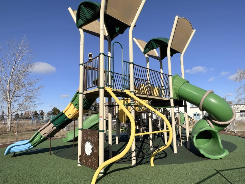 Willow Park School-14 Colorful playground with slides, climbing structures, and shades under a clear blue sky. Inviting and cheerful, set on padded green flooring.