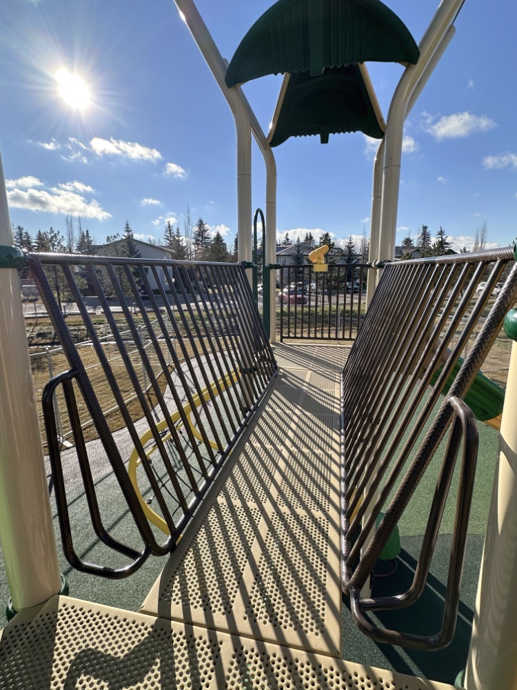 Willow Park School-15 Playground bridge walkway at Willow Park School with metal railings casts shadows, under a sunny blue sky. Surrounding trees and houses add a serene, playful tone.