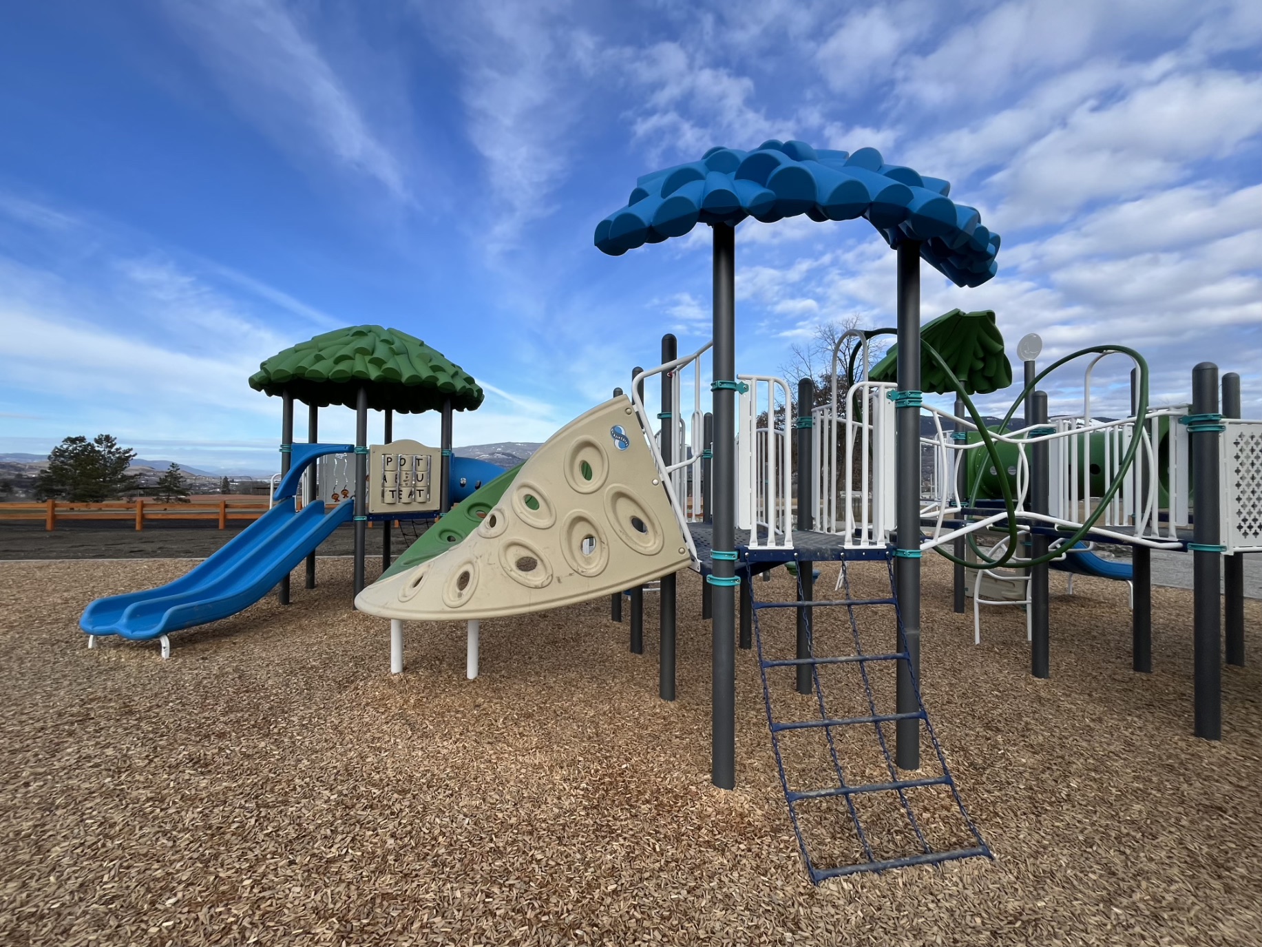 Blue slide, beige climbing wall and chain ladder under a blue leaf canopy, mountains behind.