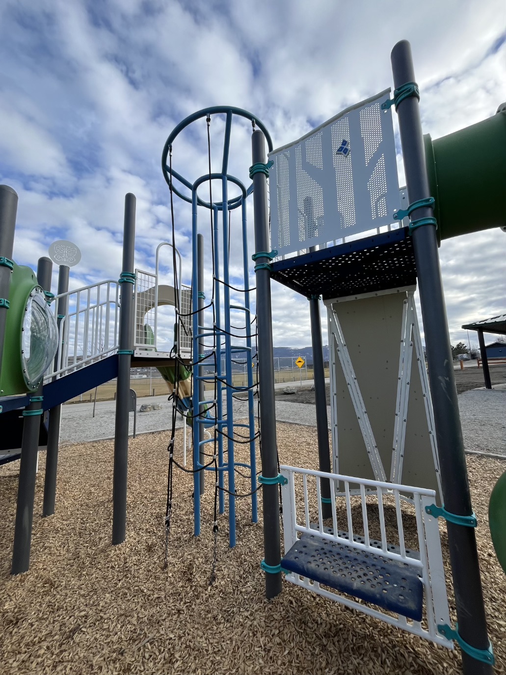 Blue rope climbing tower beside a perforated panel and a bench seat on the ground below.