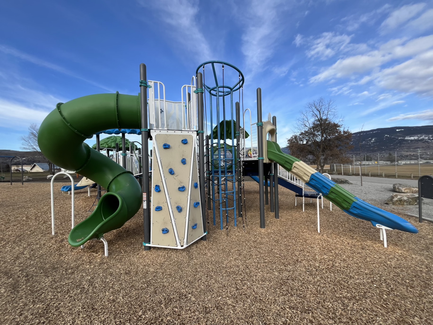 Green spiral slide and rock wall at DND Grounds Playground in Vernon, BC, with mountains behind.